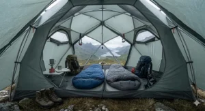 Interior view of a spacious geodesic tent with sleeping bags, rucksacks, and a camping stove neatly arranged on a groundsheet.