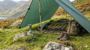 Internal view of a wild camping setup under a green tarp featuring a sleeping bag, camouflage groundsheet, and a rucksack with a Union Jack patch.