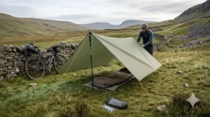 A photorealistic 4K image showing a sage-green ultralight bikepacking tarp configured as an A-frame shelter on a grassy moorland using two trekking poles.