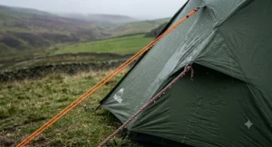 Alt text for image 5: A 4K photorealistic close-up of two distinct guy ropes under tension on a green tent flysheet in a rain-soaked Welsh mountain environment. The bright orange reflective paracord on the left remains taut and is shedding water, while the dull, twisted standard nylon rope on the right is heavily saturated, visibly sagging, and clinging with large water droplets. Diffuse natural light from a grey sky illuminates the scene.