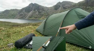 A detailed photograph showing a person in a blue knitted jumper pointing at a repaired tent pole section on a green tent. The repaired section is secured with a silver aluminium sleeve. The tent is pitched on a grassy hill overlooking a lake and mountains under a cloudy sky, with an inset close-up showing the repair sleeve in detail.