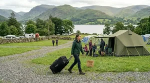 A wide illustration of a female camper pulling her wheeled tent bag across a gravel path, with other holidaymakers socialising outside their tents in the background.