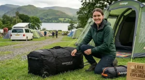 A close-up illustration of a camper checking the heavy-duty zips and durable fabric of a black wheeled tent bag on a grassy campsite pitch.