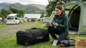 A female camper kneeling on the grass next to a packed wheeled tent bag, using a compass and map to navigate her route in the Lake District.
