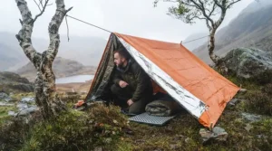 A makeshift emergency shelter constructed using the orange and silver thermal blanket and cordage in a rugged landscape.