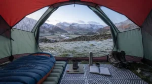 A photorealistic shot demonstrating the technique of stacking two sleeping mats inside the tent. A thick, ridged closed-cell foam mat is laid underneath a high-R-value self-inflating mat. A hand is pointing to the clear R-5.8 R-value markings on the inflatable mat, emphasising the importance of a high combined R-value for winter insulation.