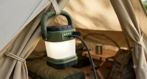 A photorealistic close-up focusing on the side of the specific olive-green tent light. It shows the detailed rugged casing and the illuminated green battery indicator LEDs, which visually demonstrate the USB-C rechargeable feature. The scene is inside a well-lit canvas tent.