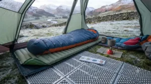 A detailed, close-up shot focusing on the tent floor, showing the layers of insulation in a cold environment. A silver-coated bubble insulation layer and a ridged foam mat are placed directly onto the frosty, icy ground beneath the tent groundsheet, illustrating the barrier against frozen earth.