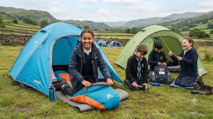 Group of diverse British primary school pupils on a residential camping trip, with one girl kneeling to pack a blue and orange junior sleeping bag into its stuff sack. Tents are pitched in a grassy field with rolling hills and dry stone walls in the background. sleeping bag for school camping trips