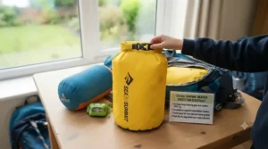 Bright yellow waterproof dry bag being sealed by a child's hand, protecting a sleeping bag and other kit inside a partially packed rucksack, positioned near a window showing a garden.