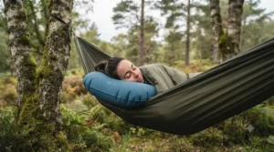 A hiker (female, with brown hair) is resting comfortably, eyes closed, in an olive-green woodland camping hammock suspended between two moss-covered birch trees in a Caledonian forest. The blue inflatable hiking pillow is properly positioned, supporting her head. The scene is bathed in soft, natural, overcast British daylight.