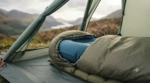 The blue inflatable hiking pillow is shown fully inflated and perfectly nested within the contoured hood of an olive-green down sleeping bag. The scene is inside a lightweight backpacking tent pitched in the Scottish Highlands, with the blurred heather-clad hills visible outside the tent opening.